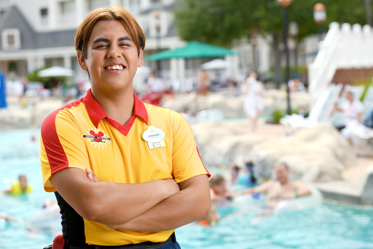 Joshua, a Disney College Program participant, in front of a pool where guests are swimming and lounging.