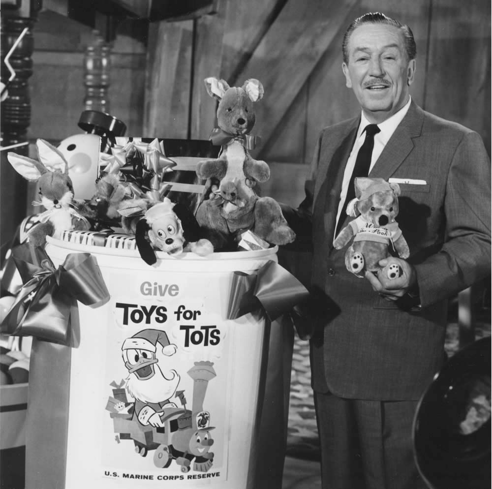 Walt Disney holding a stuffed animal next to a bin of toys reading Give Toys for Tots.
