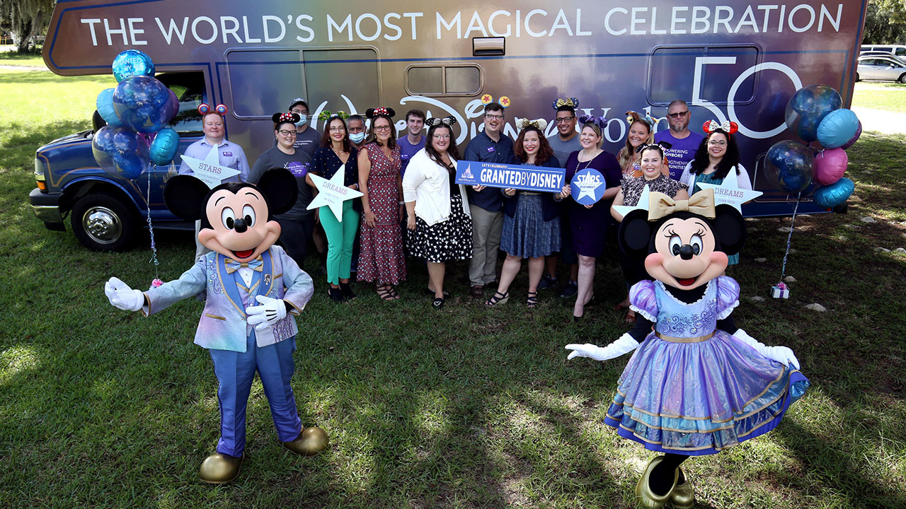 Mickey Mouse and Minnie Mouse are dressed in Walt Disney World 50th Anniversary attire and are standing in front of a shuttle bus as they join cast members during a surprise check presentation to employees of HOPE Partnership in Osceola County