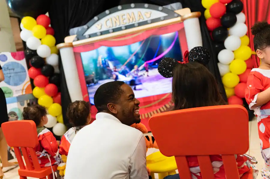 Walt Disney World Ambassador Raevon sits next to a child wearing Mickey Mouse ears enjoying the new mobile movie theater unit