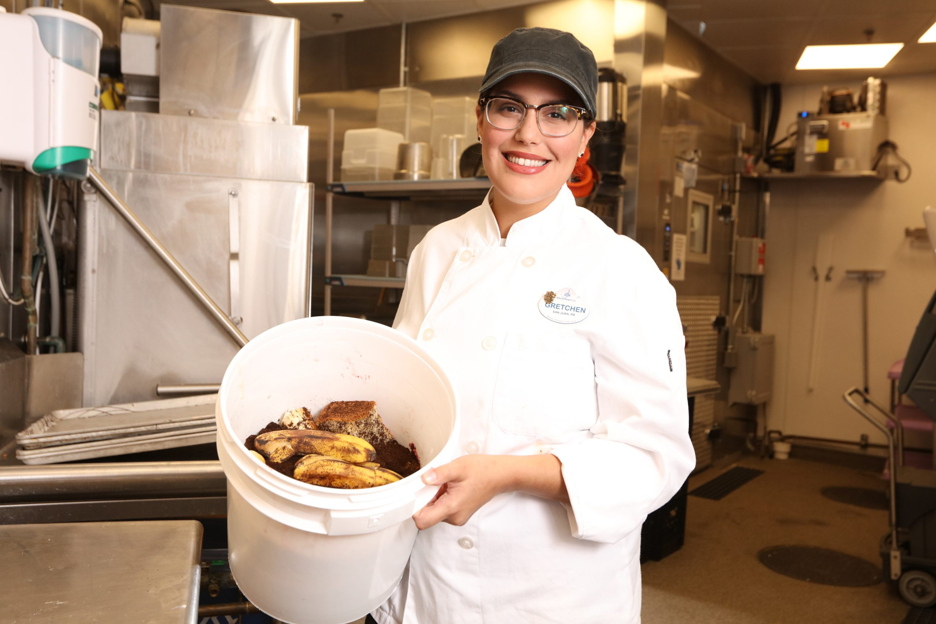 A Disney culinary cast member holding a plastic bucket of food waste in a kitchen.