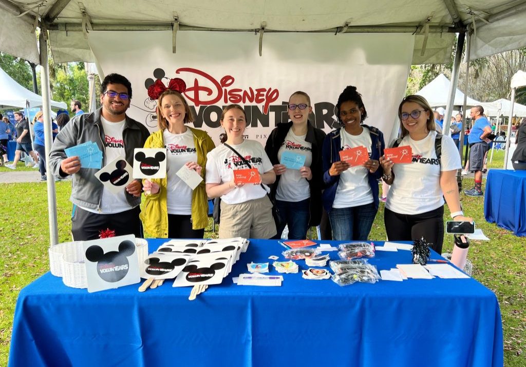 People wearing Disney VoluntEARS shirts and holding collateral while standing under a tent behind a table with a blue cover.