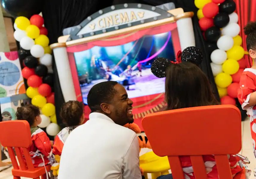 Walt Disney World Ambassador Raevon sits next to a child wearing Mickey Mouse ears enjoying the new mobile movie theater unit