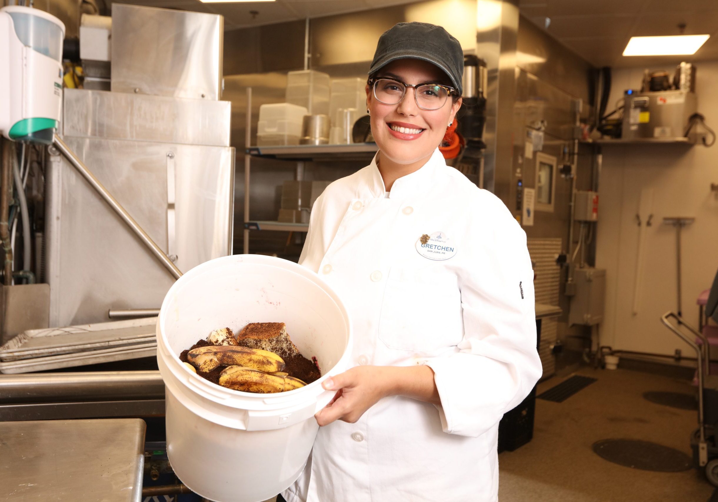 Food Waste Reduction 3 A Disney culinary cast member holding a plastic bucket of food waste in a kitchen.