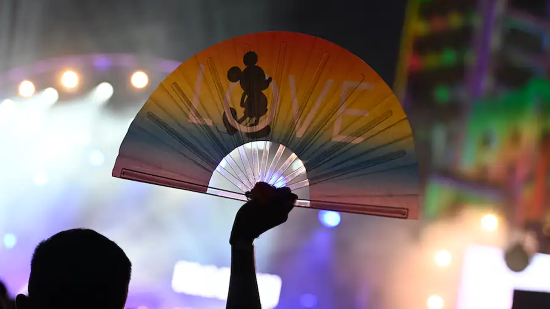 A rainbow fan is being held in the air at a concert. On the fan is a picture of Mickey and the word LOVE.