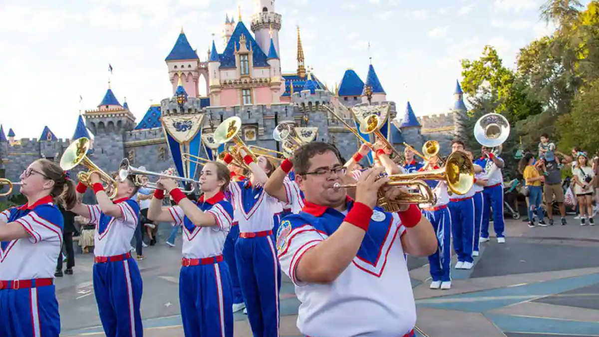 Brass members of the Disneyland All-American College Band play their instruments in front of Sleeping Beauty Castle wearing red, white, and blue uniforms.