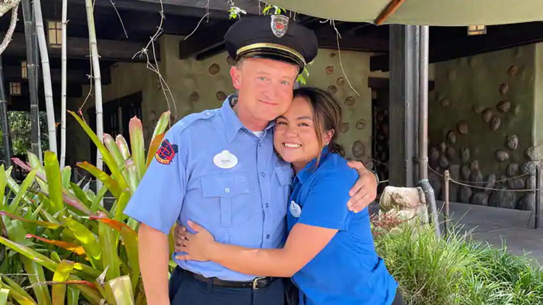 The daughter in a blue shirt wearing a nametag, hugs her dad who is wearing a Disney Security costume.