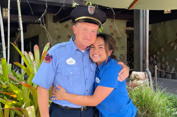 The daughter in a blue shirt wearing a nametag, hugs her dad who is wearing a Disney Security costume.