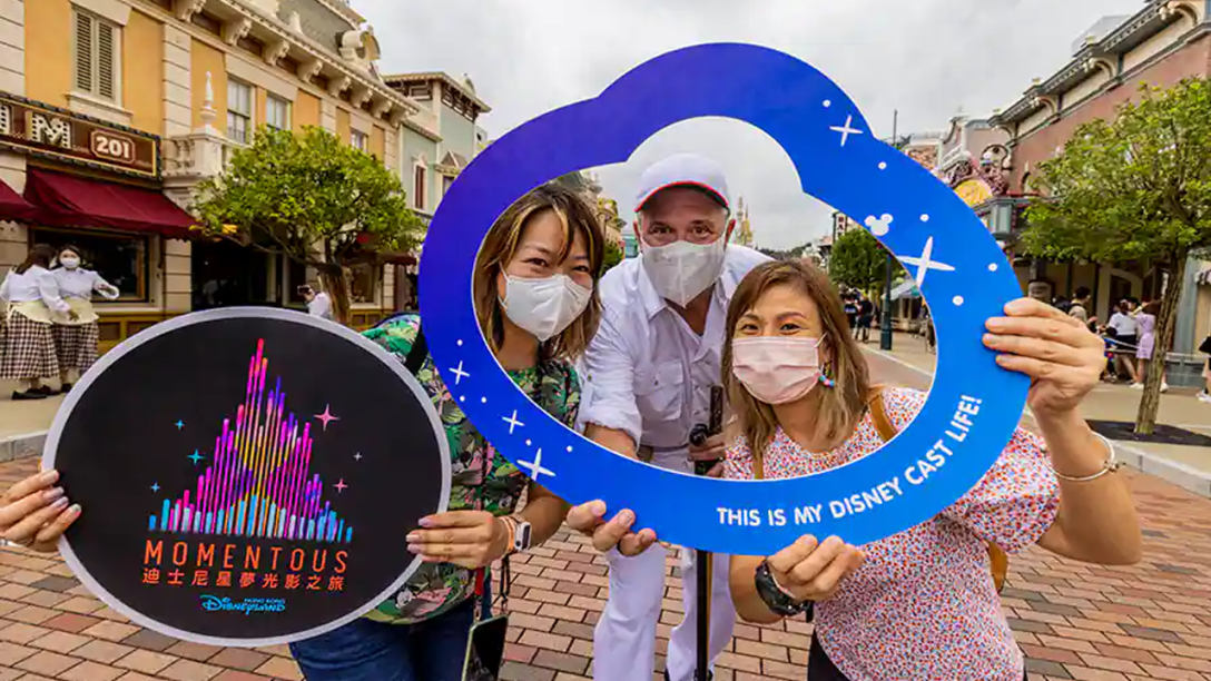 Three Hong Kong Disneyland Cast Members post holding up "Momentous" signs and frames.