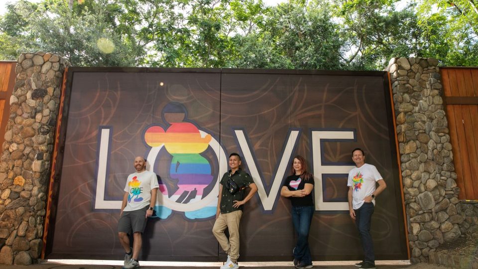 Four people standing in front of a gate with the word LOVE spelled out and a rainbow Mickey inside the O.