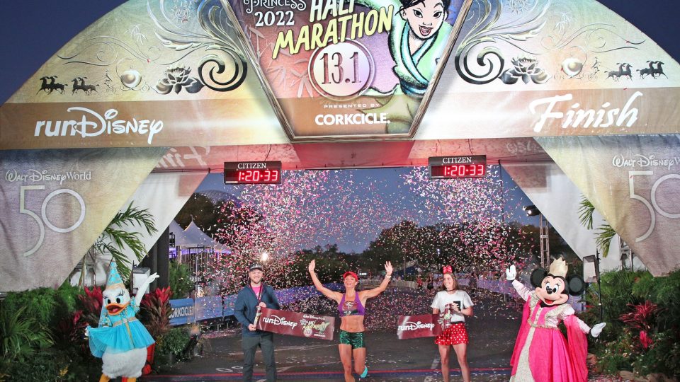 The first female runner crossing the finish line during the runDisney Princess Half Marathon with Daisy and Minnie cheering her on.