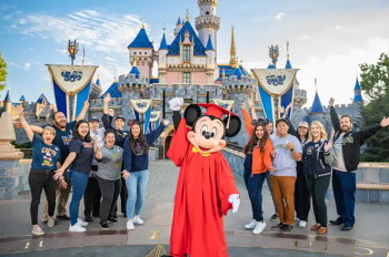Mickey wearing red graduation robes standing outside Sleeping Beauty Castle with a dozen Aspire students.