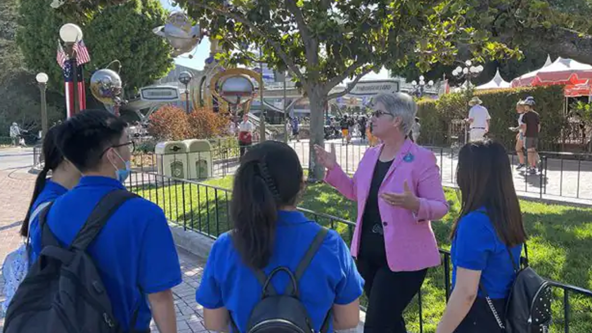 Disneyland Cast Member and Mentor is wearing a pink blazer talking to three students in royal blue tshirts.