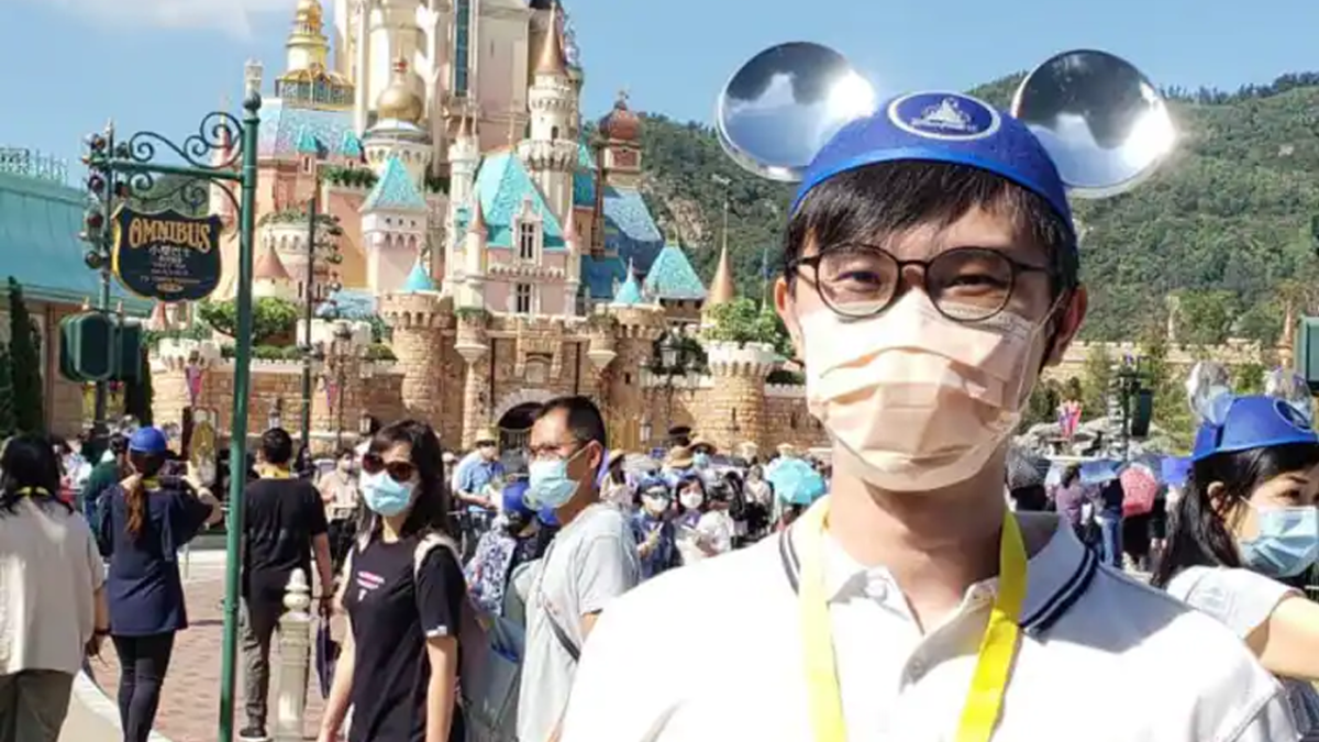 Kingsley standing in front of the castle at Hong Kong Disneyland wearing blue Mickey Mouse ears.
