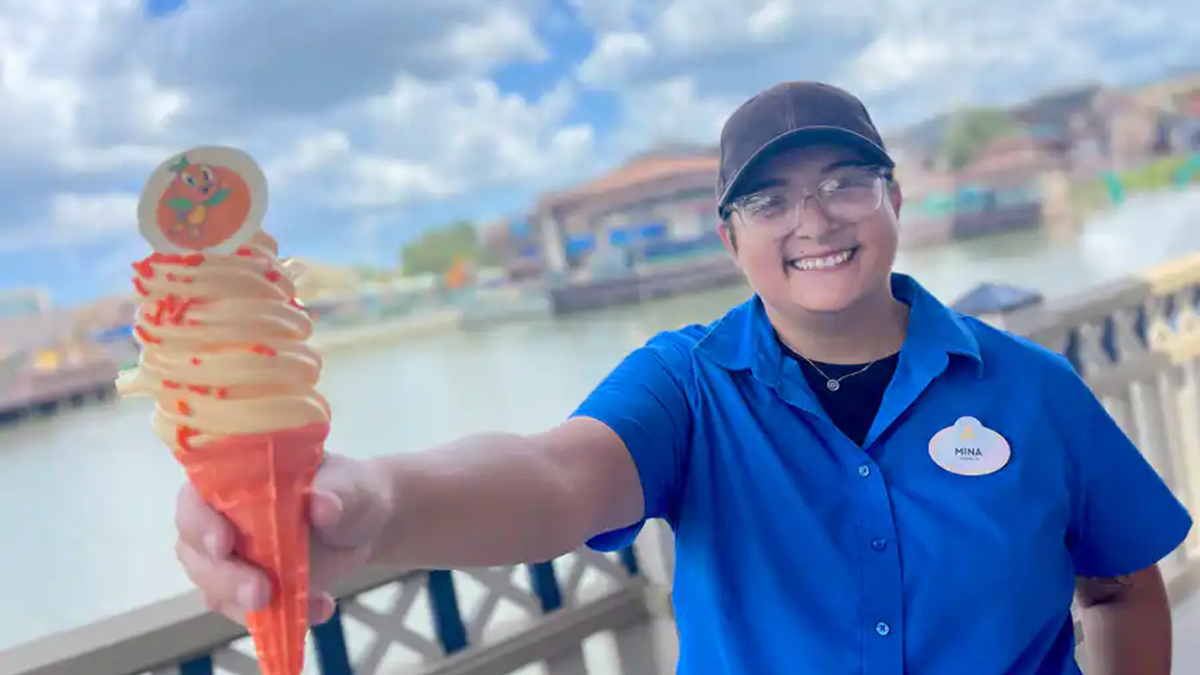 Cast Member, Mina, wearing a blue shirt and black ballcap holds out a Orange Bird themed swirl ice cream in an orange cone.