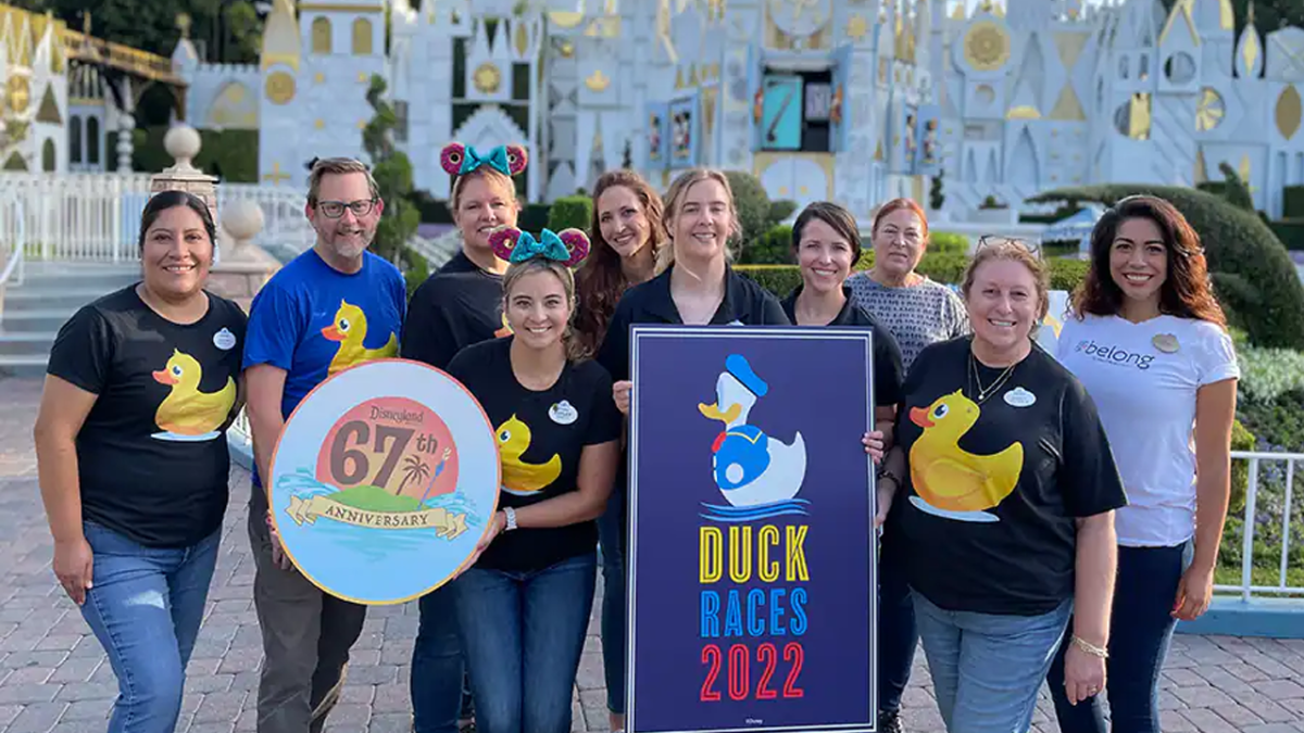 9 Cast Members wearing ducks on their shirts pose in front of it's a small world with signs that read "Duck Races 2022" and Disneyland's 67th Anniversary.
