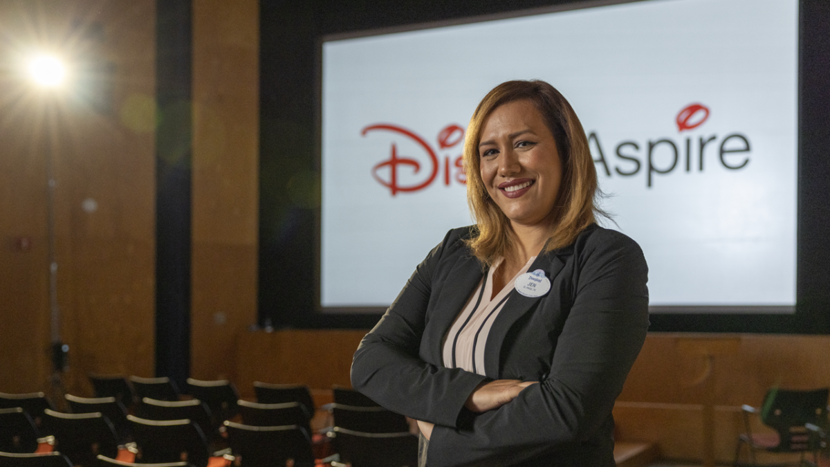 Jennifer wearing a black blazer and Disneyland nametag standing in front of a video screen that says Disney Aspire in a theater.