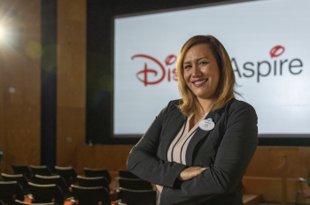Jennifer wearing a black blazer and Disneyland nametag standing in front of a video screen that says Disney Aspire in a theater.