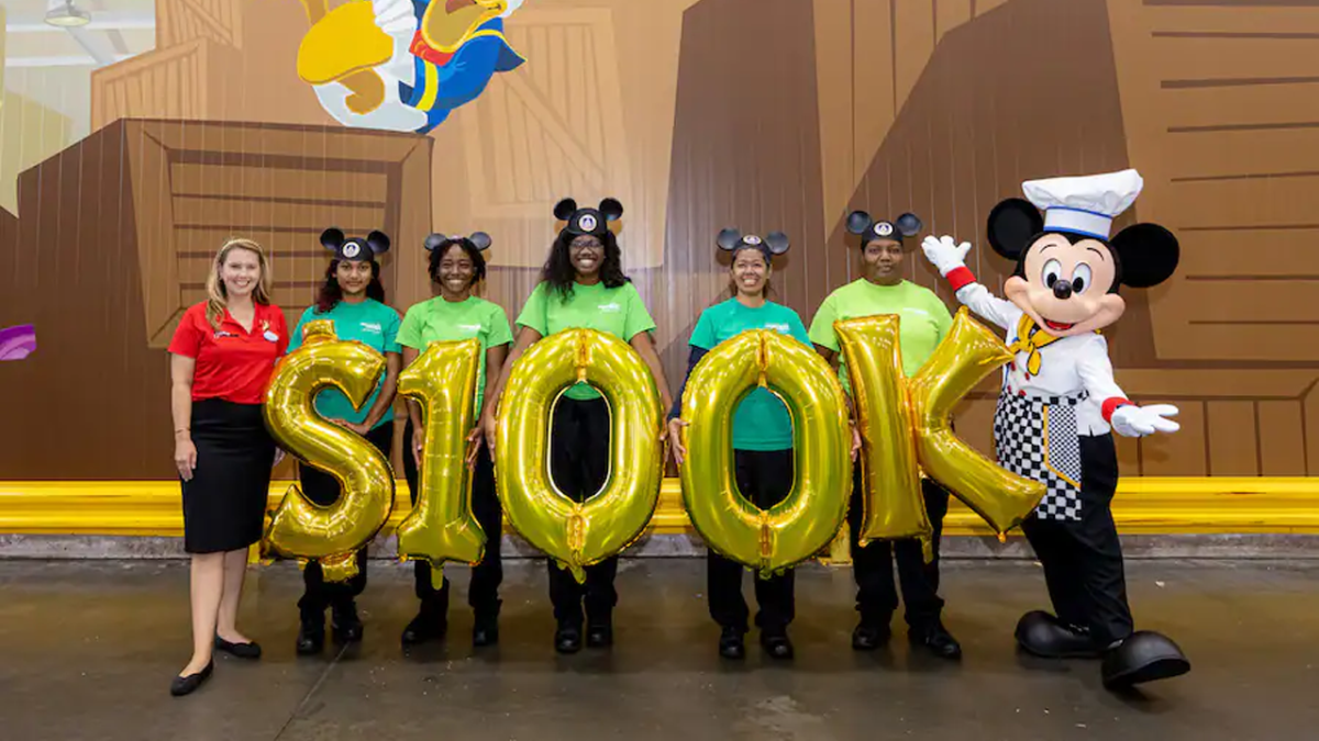 Disney Ambassador and Mickey along with five volunteers hold balloons spelling $100K.