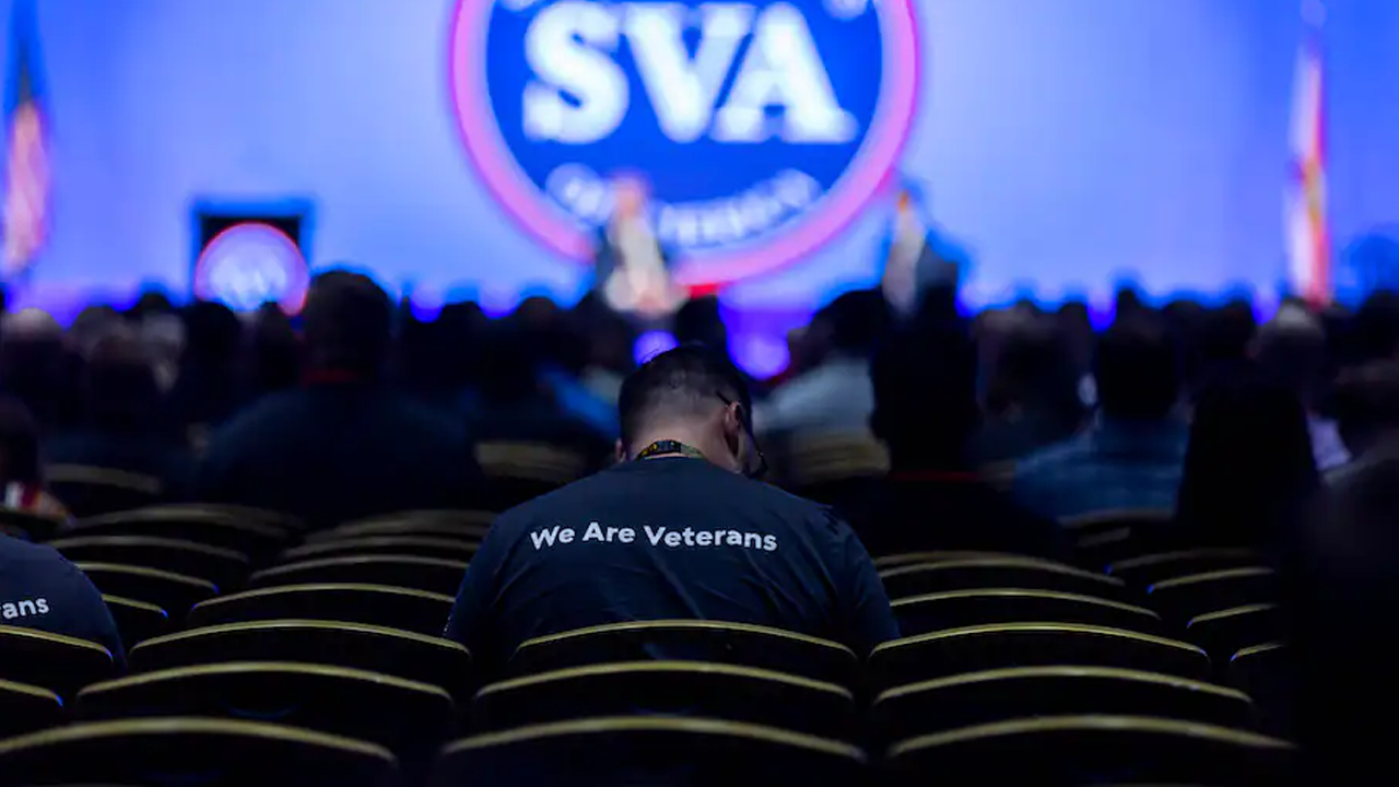 Man sitting in an auditorium. The back of his shirt says "We are Veterans."