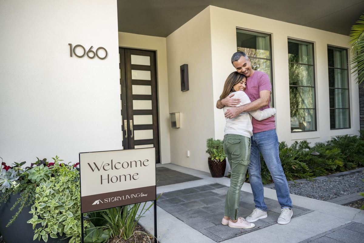 A couple hugs in front of a new Cotino home with a "welcome home" sign in front.